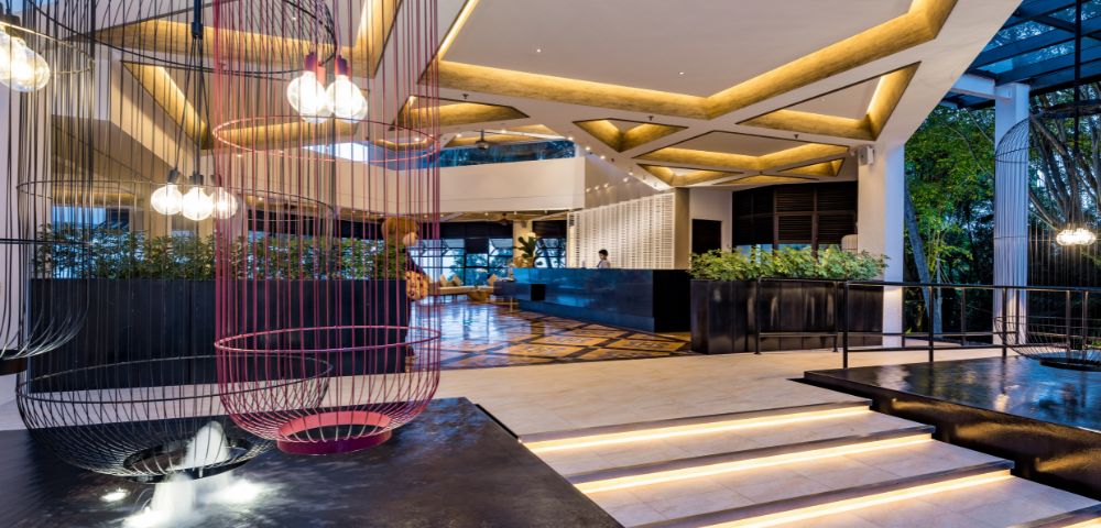 Spacious modern hotel lobby with geometric lighting, two large birdcage light fixtures, sleek white marble steps, and greenery, exuding elegance and warmth.