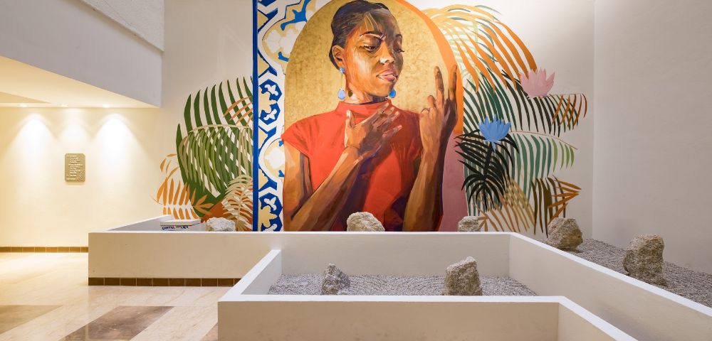 A vibrant mural depicts a thoughtful woman in a red shirt, surrounded by abstract leaves and geometric patterns. The scene is situated in a modern, serene atrium.