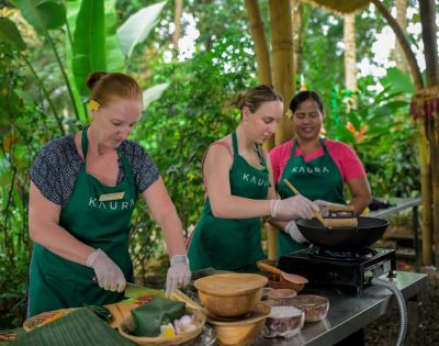 Three women wearing aprons engage in outdoor cooking with fresh ingredients on a table. The lush green backdrop creates a lively, vibrant atmosphere.