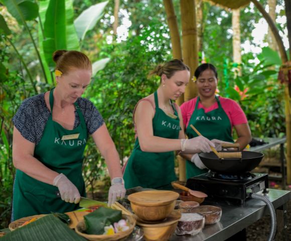 Three women wearing aprons engage in outdoor cooking with fresh ingredients on a table. The lush green backdrop creates a lively, vibrant atmosphere.
