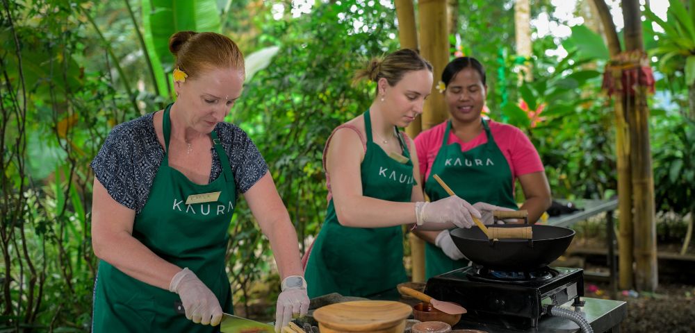 Three women wearing aprons engage in outdoor cooking with fresh ingredients on a table. The lush green backdrop creates a lively, vibrant atmosphere.