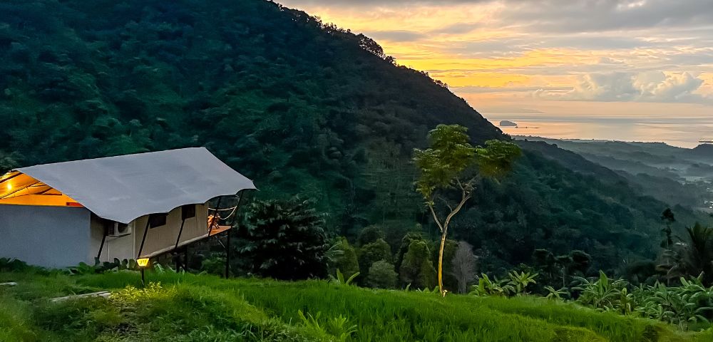 A tent perched on a lush hillside overlooks a vibrant sunset. Soft, golden light bathes the sky and sea, evoking a sense of tranquility and adventure.
