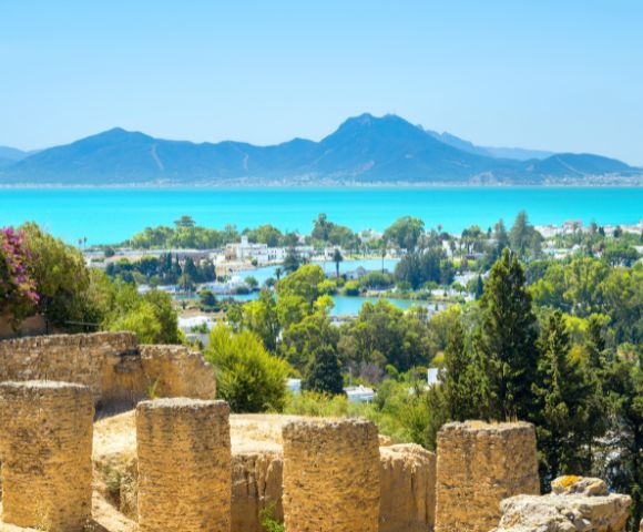 Scenic view of a coastal town with lush greenery, a sparkling blue sea, and distant mountains under a clear sky. Ancient stone ruins in the foreground.