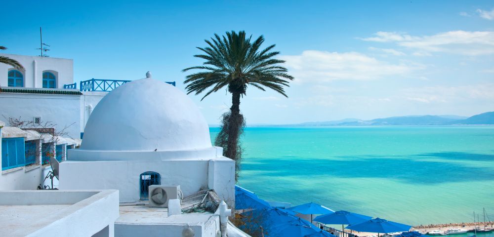 White dome-roofed buildings and palm tree overlook turquoise sea on a sunny day. Blue parasols line the shore, evoking a tranquil Mediterranean vibe.