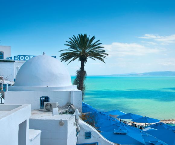 White dome-roofed buildings and palm tree overlook turquoise sea on a sunny day. Blue parasols line the shore, evoking a tranquil Mediterranean vibe.