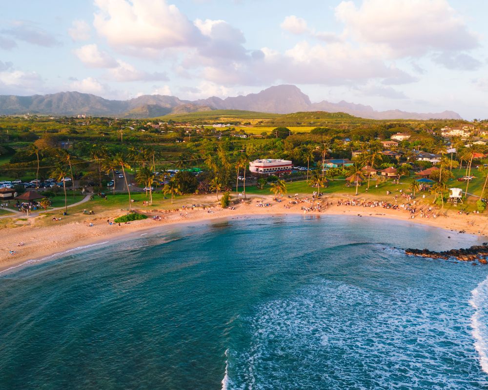 A serene beach scene at sunset with gentle waves, golden sand, and scattered beachgoers. Lush green landscape and mountains under a partly cloudy sky.