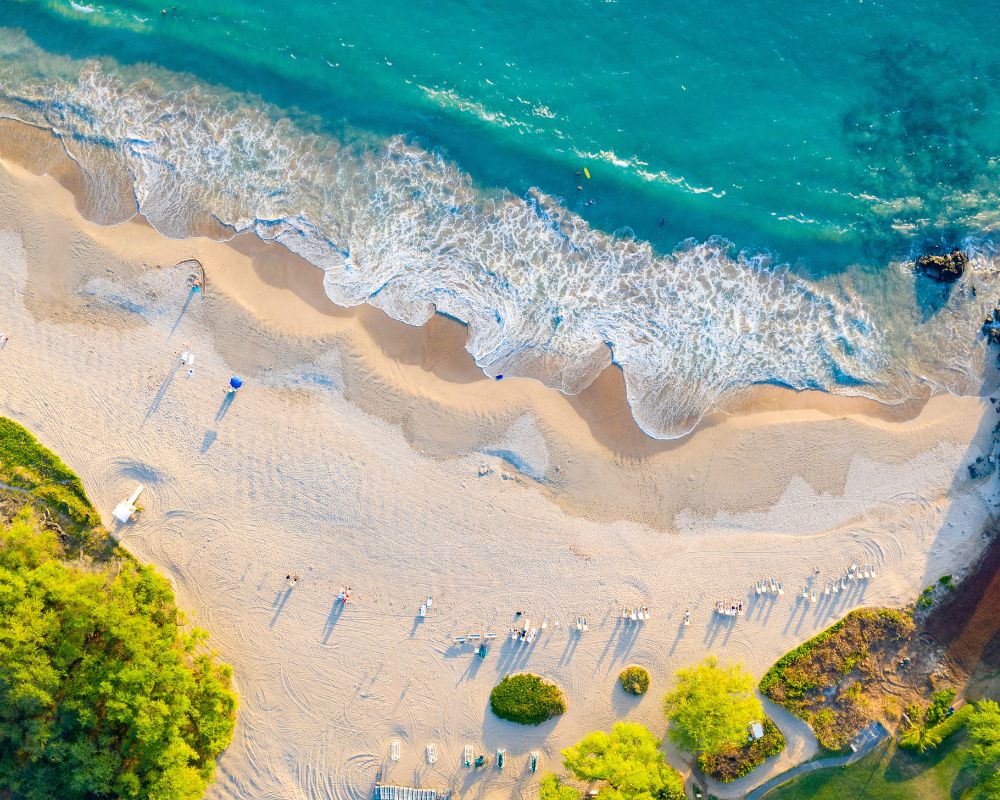 Aerial view of a sunny beach with turquoise waves gently meeting the sandy shore. People cast long shadows, conveying a relaxed and vibrant atmosphere.