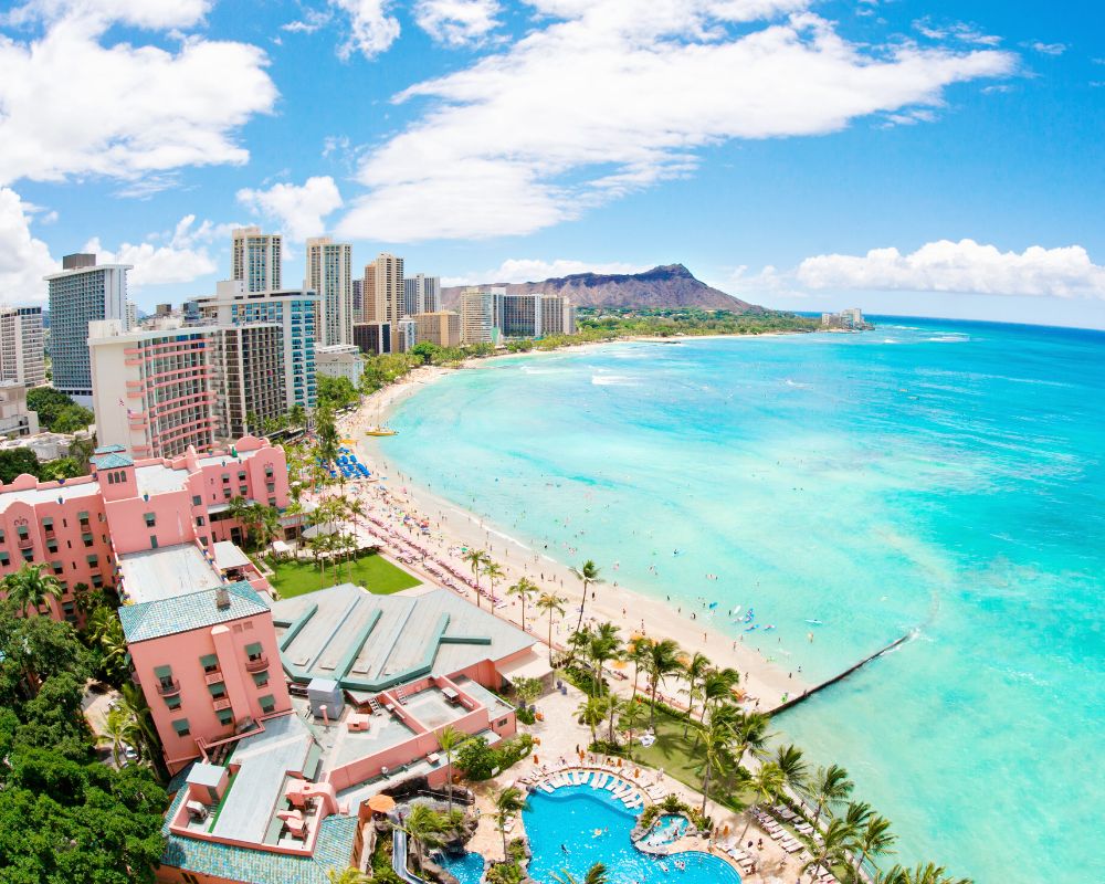 Aerial view of Waikiki Beach in Honolulu, Hawaii, with turquoise ocean waves, sandy shore, vibrant pink hotel, tall city buildings, and Diamond Head in the background.