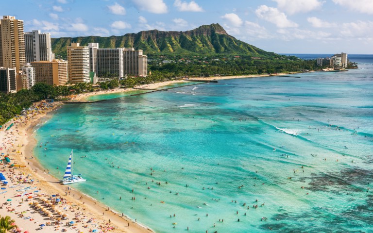 Tropical beach scene with turquoise ocean waves, crowded sandy shore, and tall city buildings. Lush green mountain under a partly cloudy sky.
