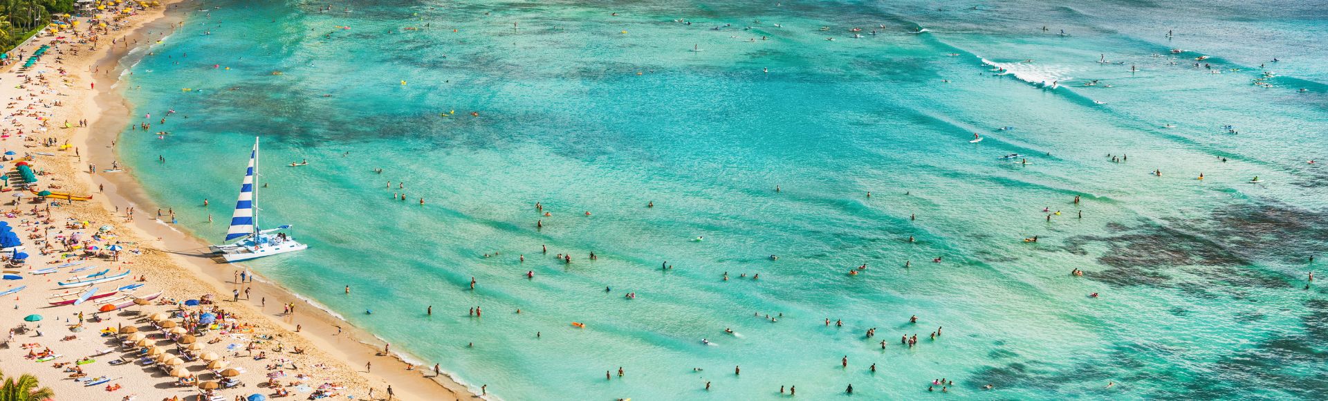 Aerial view of a crowded beach with turquoise water, swimmers, and a sailboat with blue-and-white stripes. Sandy shore lined with sunbathers and umbrellas.