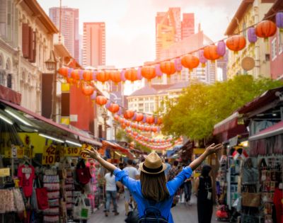 A person with a hat and backpack joyfully raises their arms in a bustling street market adorned with red lanterns and surrounded by tall city buildings at sunset.
