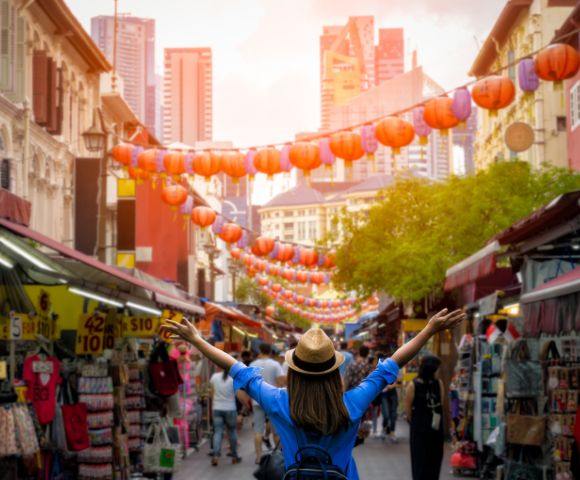 A person with a hat and backpack joyfully raises their arms in a bustling street market adorned with red lanterns and surrounded by tall city buildings at sunset.