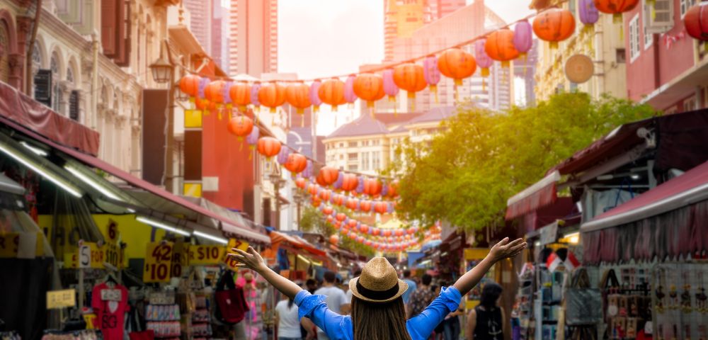 A person with a hat and backpack joyfully raises their arms in a bustling street market adorned with red lanterns and surrounded by tall city buildings at sunset.