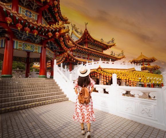 A woman in a floral dress and hat walks towards an ornate, colorful temple at sunset. The sky is orange, and the scene exudes a serene, adventurous vibe.