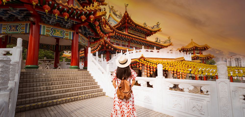 A woman in a floral dress and hat walks towards an ornate, colorful temple at sunset. The sky is orange, and the scene exudes a serene, adventurous vibe.