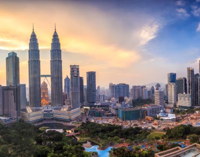 Skyline of Kuala Lumpur at sunset, featuring the Petronas Twin Towers surrounded by modern buildings. The sky is a blend of orange and blue hues.