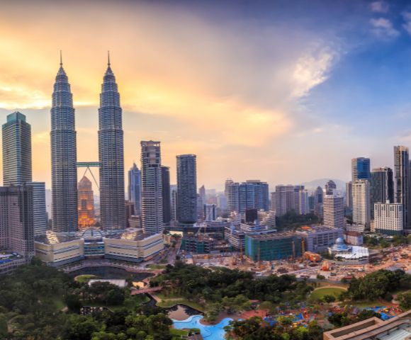 Skyline of Kuala Lumpur at sunset, featuring the Petronas Twin Towers surrounded by modern buildings. The sky is a blend of orange and blue hues.