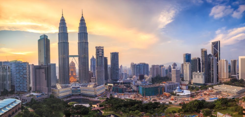 Skyline of Kuala Lumpur at sunset, featuring the Petronas Twin Towers surrounded by modern buildings. The sky is a blend of orange and blue hues.