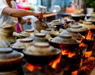 A person tends to multiple clay pots on open flames, each covered with a lid, in a bustling street food setting, conveying warmth and culinary artistry.
