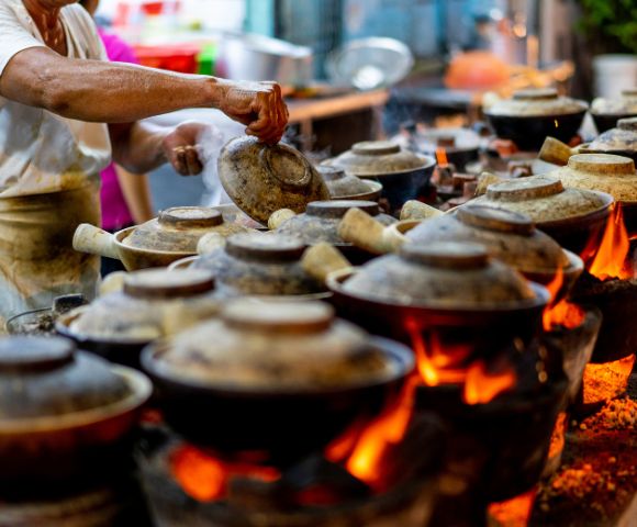 A person tends to multiple clay pots on open flames, each covered with a lid, in a bustling street food setting, conveying warmth and culinary artistry.