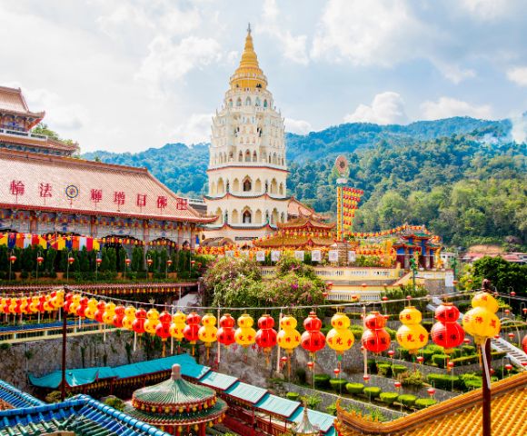 Scenic view of a vibrant Chinese temple complex with a towering pagoda, colorful lanterns, ornate rooftops, and lush green mountains in the background.