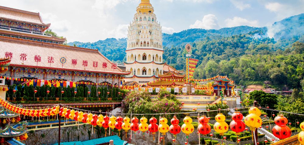 Scenic view of a vibrant Chinese temple complex with a towering pagoda, colorful lanterns, ornate rooftops, and lush green mountains in the background.
