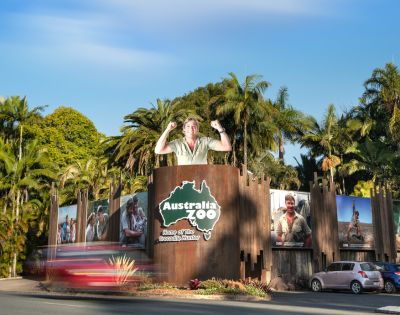 Entrance to Australia Zoo surrounded by palm trees with large wildlife posters. A statue of a person stands atop the gate, conveying excitement.