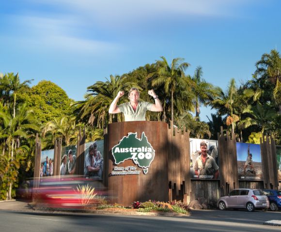 Entrance to Australia Zoo surrounded by palm trees with large wildlife posters. A statue of a person stands atop the gate, conveying excitement.
