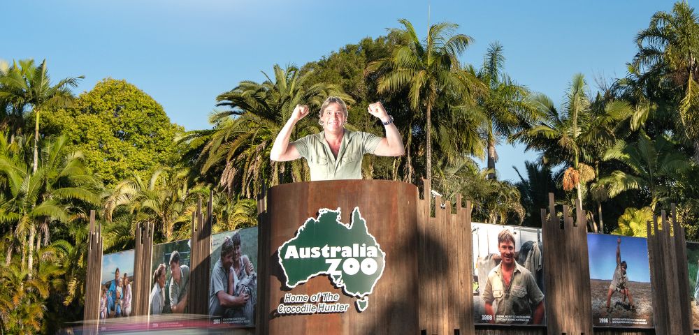 Entrance to Australia Zoo surrounded by palm trees with large wildlife posters. A statue of a person stands atop the gate, conveying excitement.
