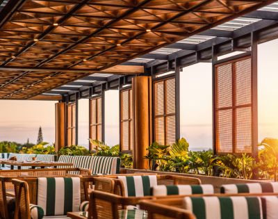 A sunlit patio with wooden lattice ceiling, striped cushions on chairs, and lush greenery. The warm, inviting atmosphere overlooks a serene sunset.