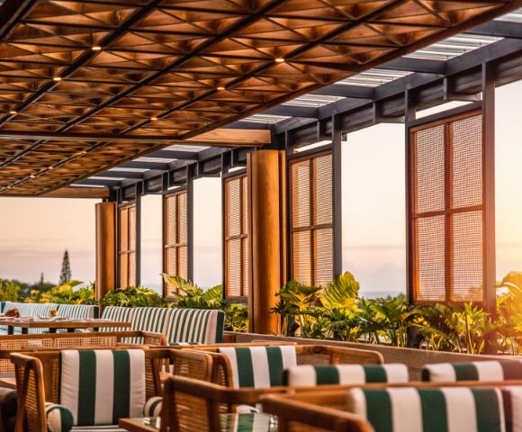 A sunlit patio with wooden lattice ceiling, striped cushions on chairs, and lush greenery. The warm, inviting atmosphere overlooks a serene sunset.