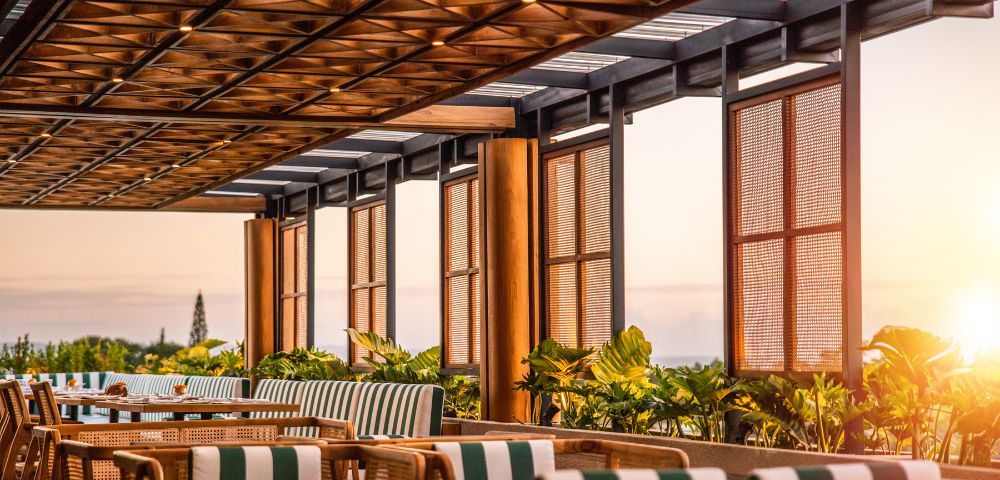 A sunlit patio with wooden lattice ceiling, striped cushions on chairs, and lush greenery. The warm, inviting atmosphere overlooks a serene sunset.