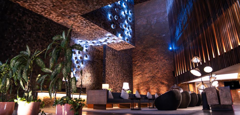 Modern lounge with textured brick walls, soft ambient blue lighting, and cozy seating. Indoor plants add a touch of nature. Relaxed, elegant atmosphere.