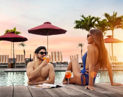 A couple enjoying drinks by a pool at sunset. They wear sunglasses and smile, surrounded by palm trees and red umbrellas, creating a relaxed tropical vibe.