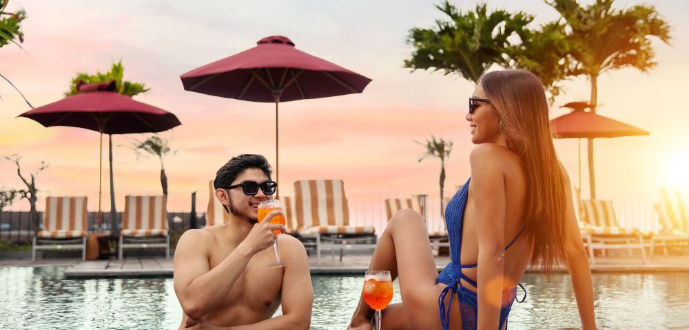 A couple enjoying drinks by a pool at sunset. They wear sunglasses and smile, surrounded by palm trees and red umbrellas, creating a relaxed tropical vibe.