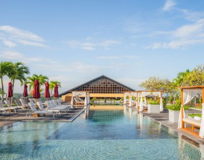 Luxurious resort pool area with clear water, lined with white cabanas, lounge chairs, and palm trees under a clear blue sky, exuding relaxation.