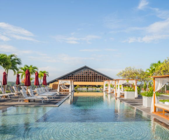 Luxurious resort pool area with clear water, lined with white cabanas, lounge chairs, and palm trees under a clear blue sky, exuding relaxation.