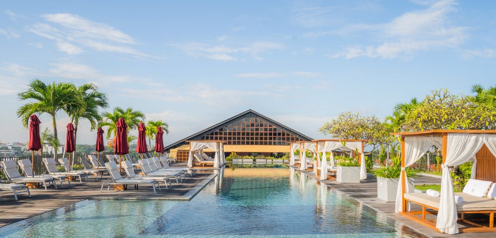 Elegant poolside cabanas with white drapes and lush greenery stand beside a reflective pool under a clear blue sky, evoking a serene, tropical paradise.