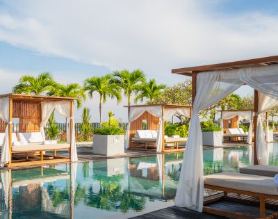 Luxurious poolside with wooden cabanas, white curtains, and lounge chairs reflecting in the water. Tropical palm trees and a bright sky enhance tranquility.