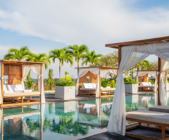 Luxurious poolside with wooden cabanas, white curtains, and lounge chairs reflecting in the water. Tropical palm trees and a bright sky enhance tranquility.