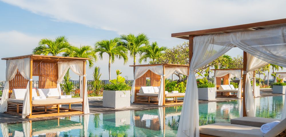 Luxurious poolside with wooden cabanas, white curtains, and lounge chairs reflecting in the water. Tropical palm trees and a bright sky enhance tranquility.