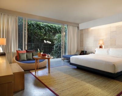 Modern hotel room with large bed, nightstand, and lamp. Seating area with a round table and couch. Floor-to-ceiling window shows lush greenery. Cozy and inviting atmosphere.