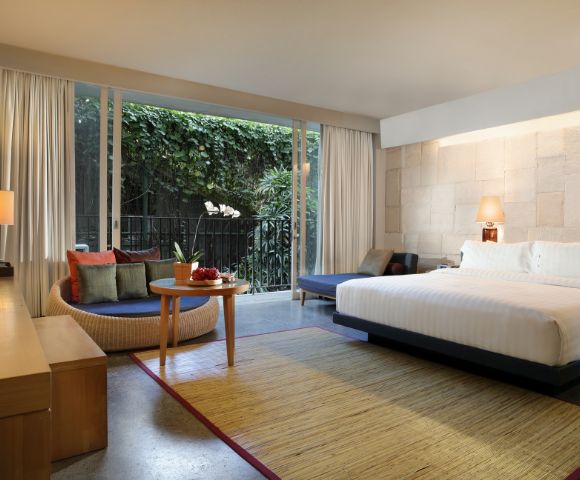 Modern hotel room with large bed, nightstand, and lamp. Seating area with a round table and couch. Floor-to-ceiling window shows lush greenery. Cozy and inviting atmosphere.