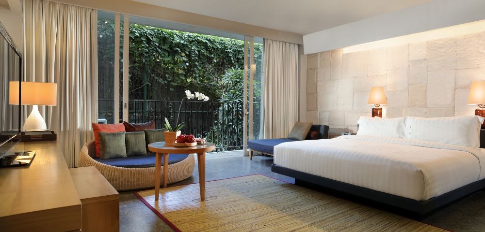 Modern hotel room with large bed, nightstand, and lamp. Seating area with a round table and couch. Floor-to-ceiling window shows lush greenery. Cozy and inviting atmosphere.