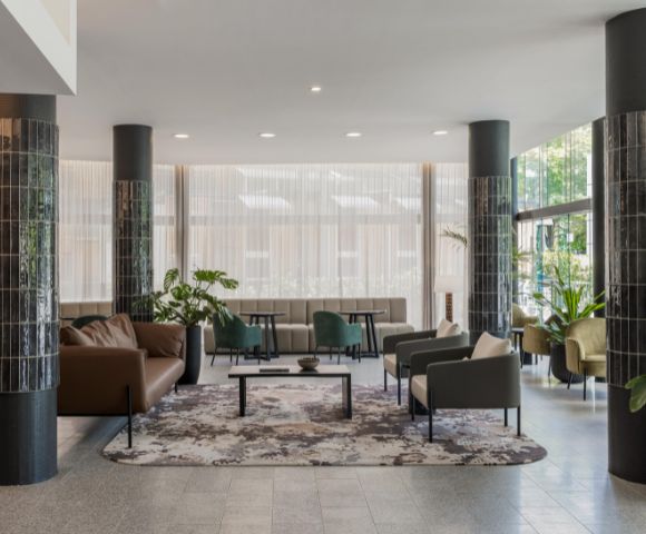 Modern lobby with sleek black columns and elegant seating. Neutral-toned sofas and armchairs surround a decorative rug. Large windows bring in natural light.