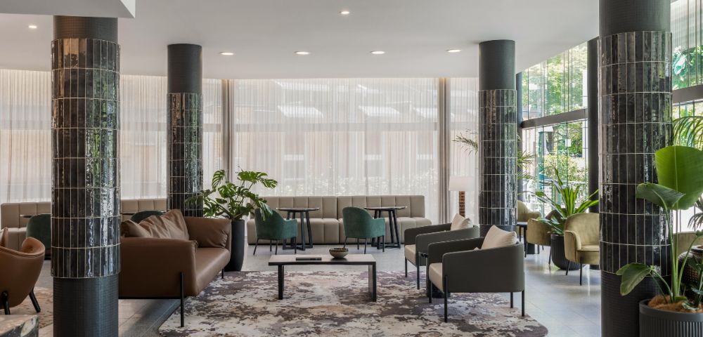 Modern lobby with sleek black columns and elegant seating. Neutral-toned sofas and armchairs surround a decorative rug. Large windows bring in natural light.