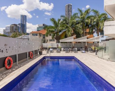 Rooftop pool scene with a bright blue water, palm trees, and city skyline. Two red lifebuoys on the wall; white lounge chairs under canopy. Sunny, relaxed vibe.