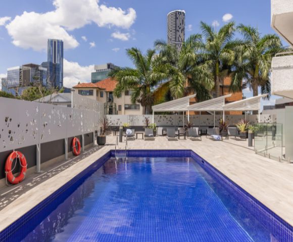 Rooftop pool scene with a bright blue water, palm trees, and city skyline. Two red lifebuoys on the wall; white lounge chairs under canopy. Sunny, relaxed vibe.