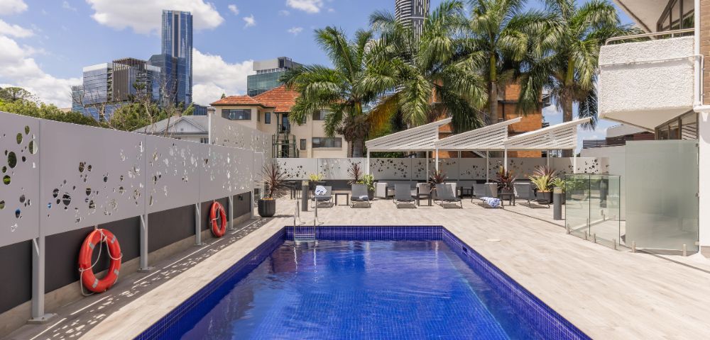 Rooftop pool scene with a bright blue water, palm trees, and city skyline. Two red lifebuoys on the wall; white lounge chairs under canopy. Sunny, relaxed vibe.