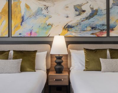 Two neatly made beds with white linens and green pillows flank a wooden nightstand with a black lamp. Abstract art with blue and yellow splashes hangs above.